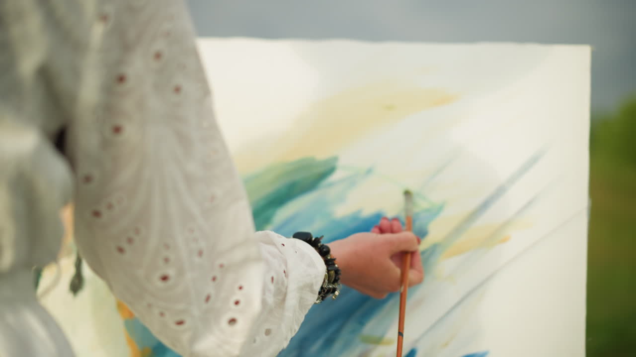 un primer plano de la mano de una mujer, adornada con cuentas, pintando delicadamente con un pincel en una paleta colorida. el artista lleva un vestido blanco, y la escena está bañada en luz suave y natural