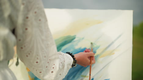 un primer plano de la mano de una mujer, adornada con cuentas, pintando delicadamente con un pincel en una paleta colorida. el artista lleva un vestido blanco, y la escena está bañada en luz suave y natural