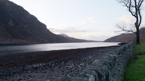 Un-Tiro-Panorámico-Lento-Se-Aleja-De-Una-Casa-Antigua-En-Las-Tierras-Altas-Escocesas-Hacia-Las-Montañas-Mientras-El-Sol-Se-Pone-En-Un-Lago-Marino