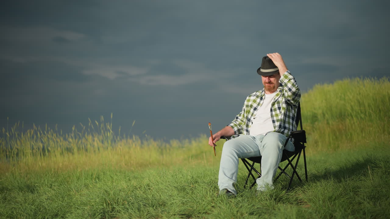 어두운 하늘 아래의 잔디에서, 체크 셔츠와 청바지를 입은 남자가 의자에 편안하게 앉아 있습니다. 그는 한 손으로 모자를 조정하면서 다른 손으로 페인트 브러쉬를 들고 있습니다.
