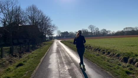 Following-shot-of-sporty-girl-running-german-marathon-outdoors-between-rural-fields-and-sunlight