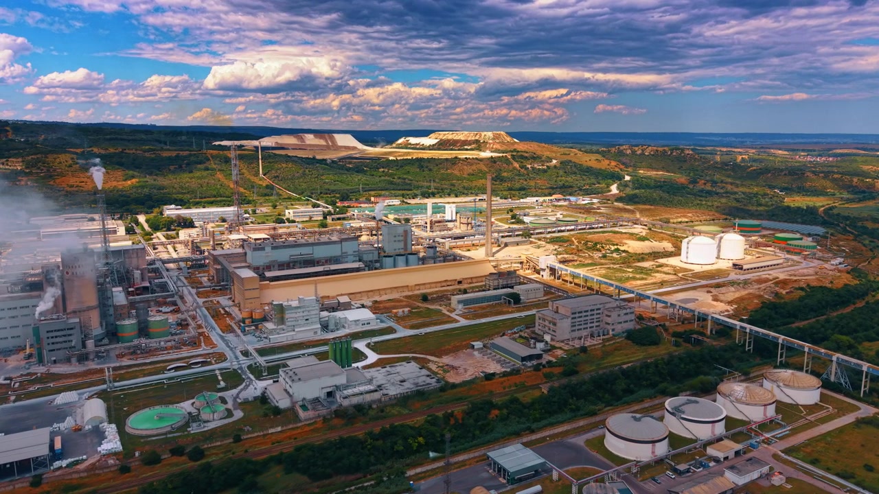 Aerial panorama of industrial processing plant under layered clouds. Storage tanks, pipelines, factory buildings and steam stacks extend across a rural valley.