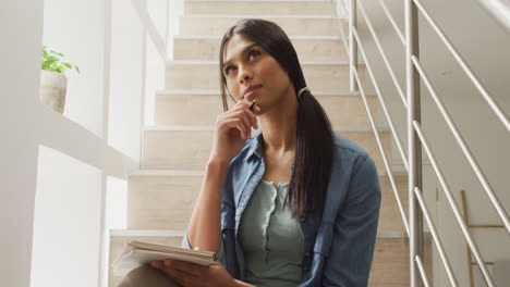 video of thoughtful biracial woman sitting on stairs and making notes