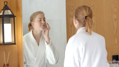 woman in bathrobe applying skincare product while looking in bathroom mirror