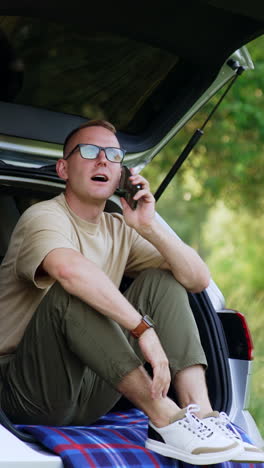 relaxed caucasian man holding smartphone. man starts to talk on the phone sitting in the car trunk. forest at backdrop. vertical video