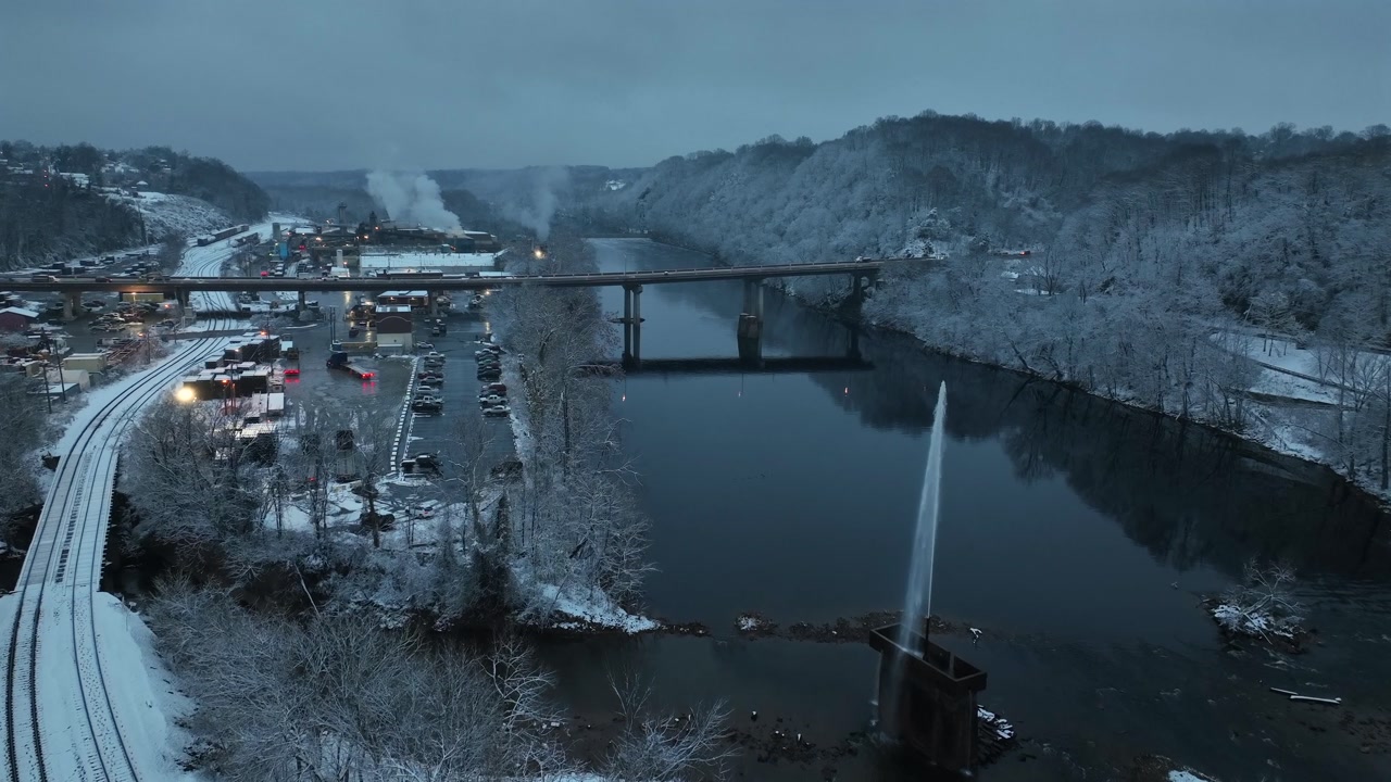 Snowy aerial view of river and fountain in Lynchburg, showing winter landscape with bridge, calm water, snow-covered hills, rail lines and moody blue dusk atmosphere. Wide shot.