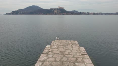 Seagulls-fly-away-from-boardwalk-of-Arona-lake-pier-with-castle-in-background