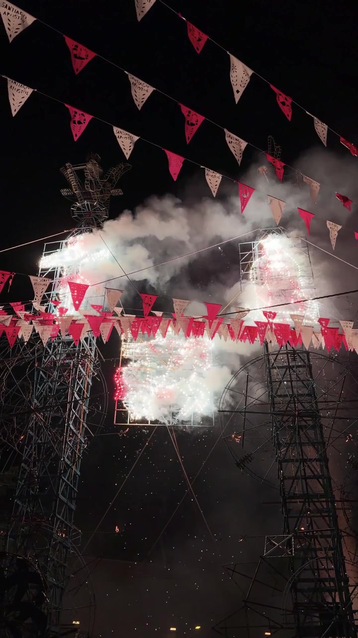 Colorful fireworks erupt from a scaffold castle at night, framed by festival bunting