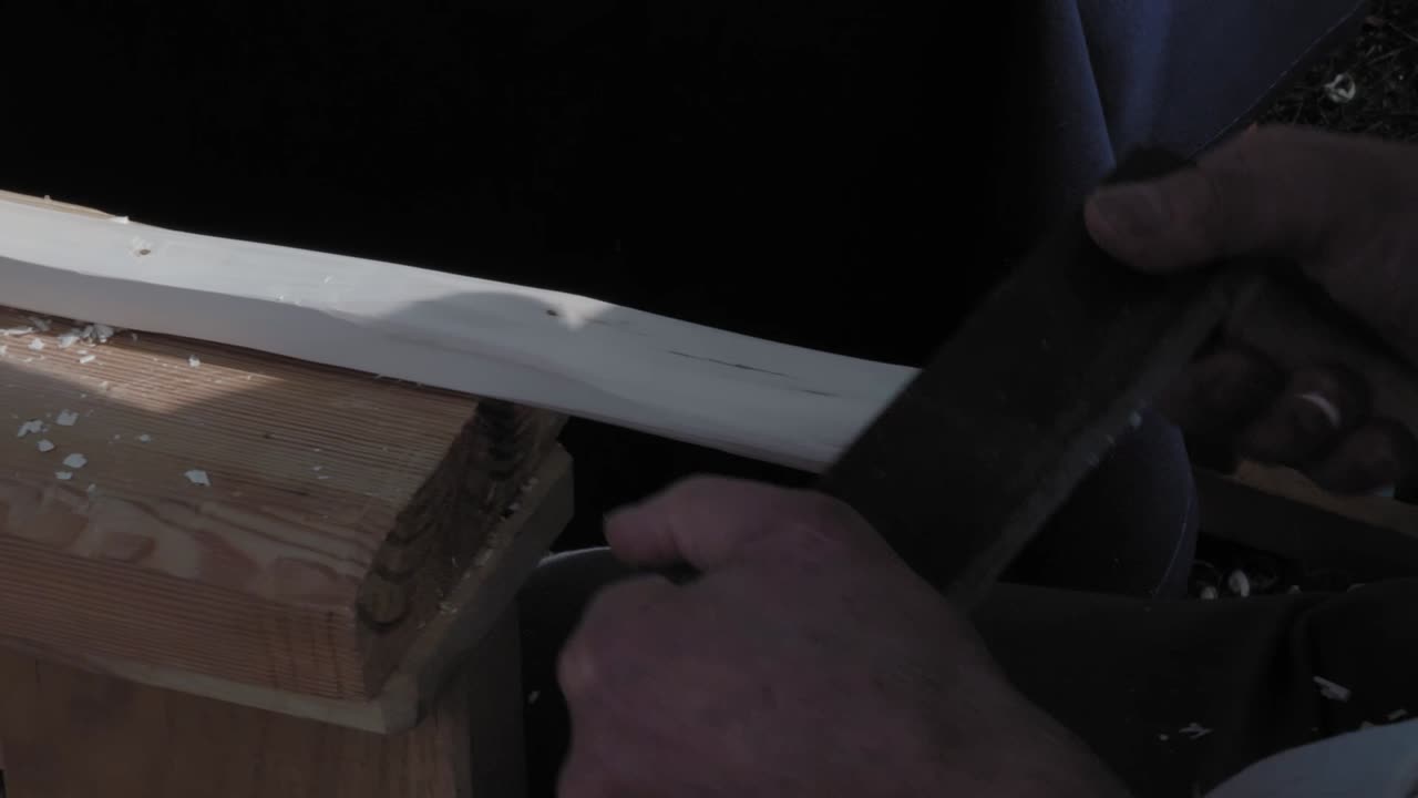 A traditional carpenter uses a metal planer to peel wood. Chips are flying. He works outside during the event. People pass by. Close-up of hands.