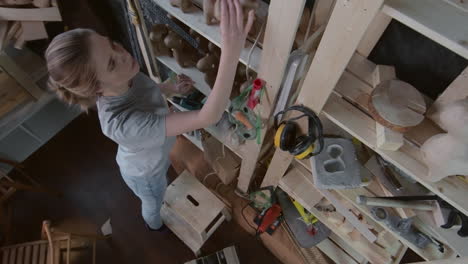 Top-View-Of-Two-Artisan-Women-In-A-Craft-Workshop,-One-Of-Them-Picks-Up-An-Item-On-The-Shelf-And-Sits-At-The-Table-With-Her-Colleague