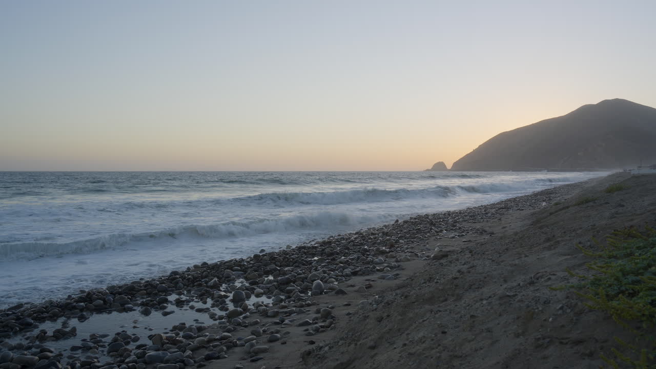 toma estacionaria de olas rodando hacia las orillas de la playa de mondo con puesta de sol detrás de la montaña en el fondo ubicada en el sur de california