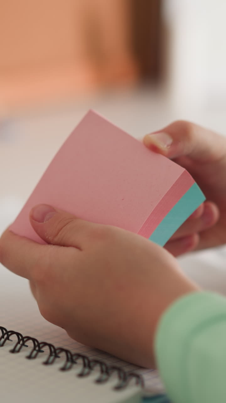niña pequeña da la vuelta a los papeles pegajosos de color en un paquete sentada en el escritorio de primer plano. niña de escuela juega con pegatinas de nota en la mesa en el lugar de trabajo de casa. suministros escolares