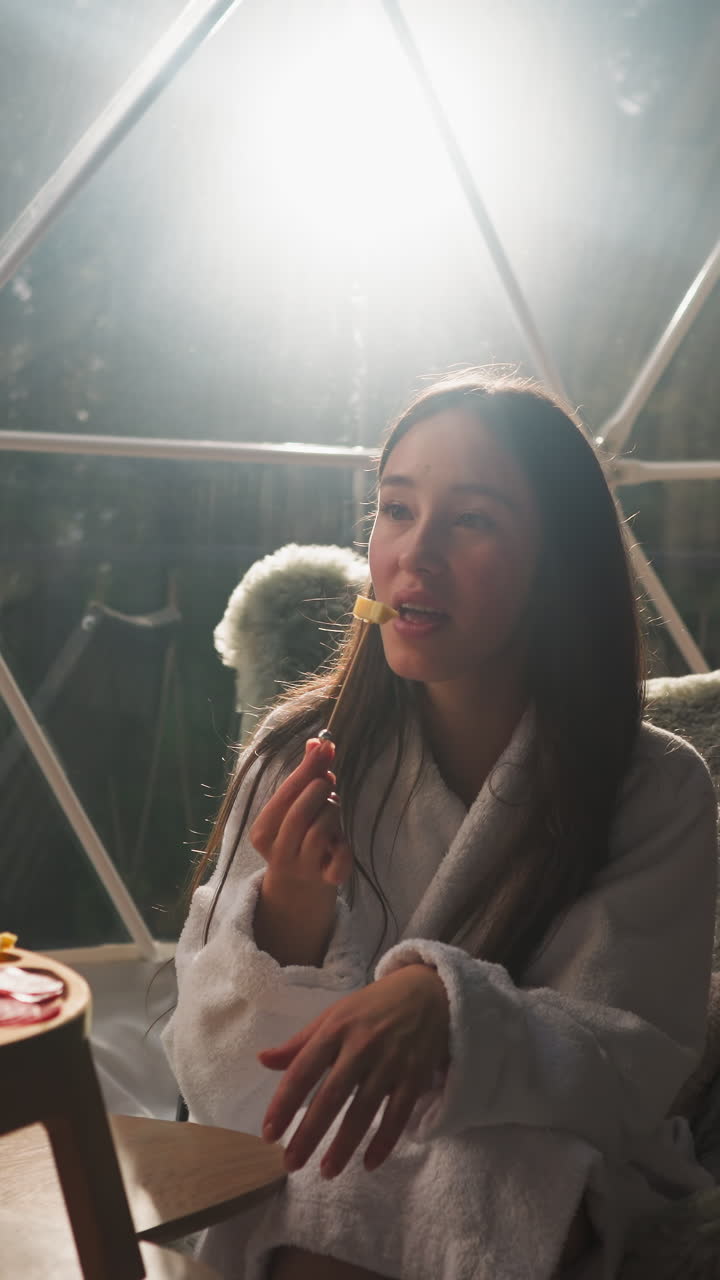 Woman eats cheese on skewer on glass terrace. Relaxed lady in terry bathrobe enjoys tasty snack sitting by window. Romantic evening with glass of wine