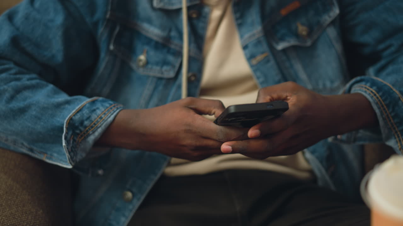 Close up of young man's hands holding smartphone while browsing or texting, dressed in denim jacket layered over hoodie, seated in cushioned chair indoors, relaxed posture, warm lighting