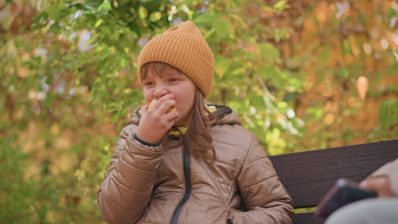 young girl in warm coat and beanie holding bitten apple and smiling warmly while sitting on bench during golden autumn, partial view of adult nearby observing child