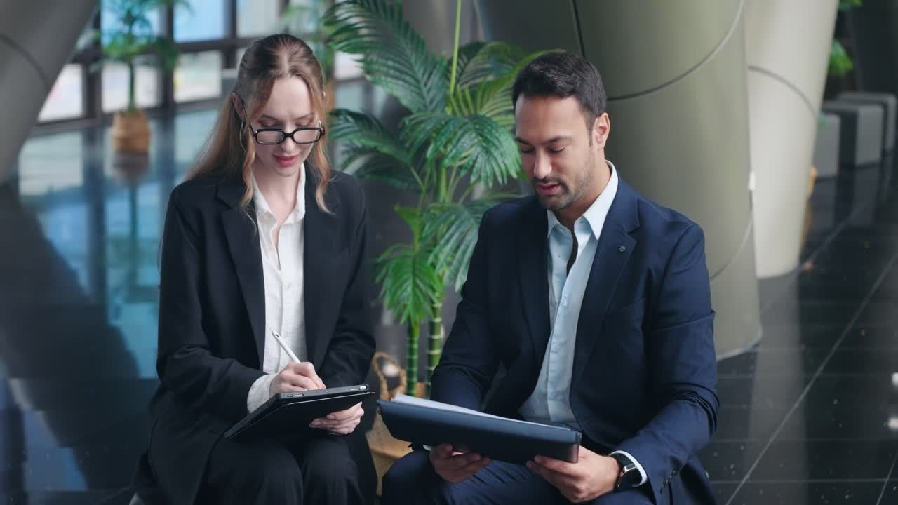 Experienced businessman sits with female employee giving constructive feedback while she takes notes on digital tablet in modern corporate office lobby during meeting