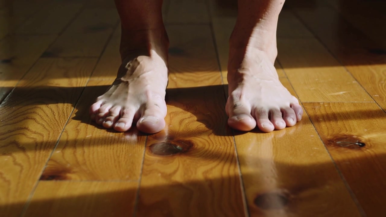 Spostando il peso, a piedi nudi che spingono in avanti bilanciandosi a casa su un pavimento di legno con una cavigliera e raggi di sole.