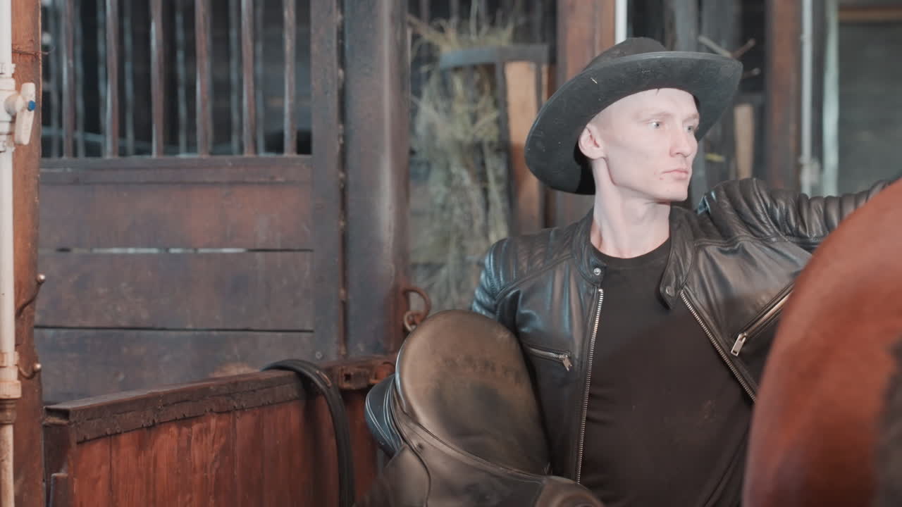 Hvid mand sadler hest i stald, fokuseret træner demonstrerer korrekt sadelplacering og justering af udstyr, læderjakke og cowboyhat, rolig hest, praktisk undervisning og samarbejde.