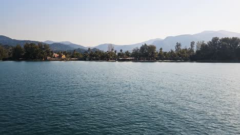 Sunlit-tropical-coastline-of-Koh-Chang-with-gentle-waves-and-a-clear-sky,-framed-by-lush-greenery-and-mountains