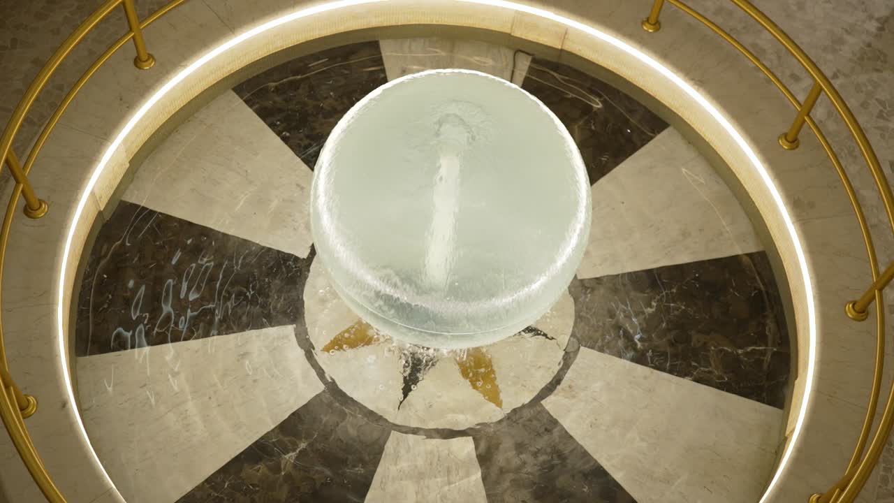 Una fontana si trova al centro di un'area circolare. La fontana presenta una ciotola di vetro da cui scende l'acqua. Il marmo circostante aggiunge un tocco di eleganza allo spazio.