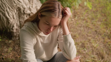 mujer joven sentada en el suelo con la cabeza apoyada en la mano izquierda y la mano derecha en la rodilla, apareciendo profundamente perdida en el pensamiento, con un fondo de corteza de árbol y vegetación