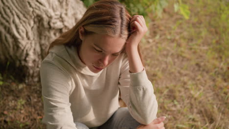 young woman seated on ground with head resting on left hand and right hand on knee, appearing deeply lost in though, with a background of tree bark and greenery