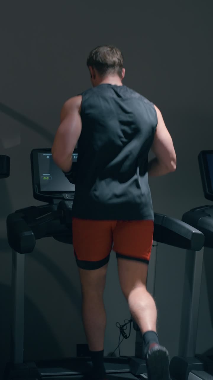 Uomo caucasico muscoloso sulla trentina corre ad alta velocità su un tapis roulant per un allenamento cardio ad alta intensità, ripreso da dietro in una palestra moderna e buia