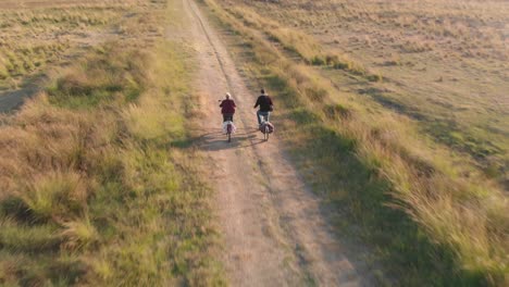 aerial drone shot of two people riding a bike on a dirt road in the middle of heathland moving from a backlit side view to behind shot revealing the wider surroundings and horizon