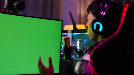 close up back view of asian boy streamer playing game over network on green screen computer. challenge and bully competitor, live stream video game, desk illuminated by rgb led strip light