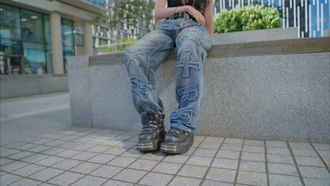 Close-Up-Outdoor-Fashion-Shot-Showing-Jeans-And-Boots-Of-Young-Alternative-Style-Woman-With-Tattoos-Sitting-Outside-Modern-Buildings-Shot-In-Real-Time