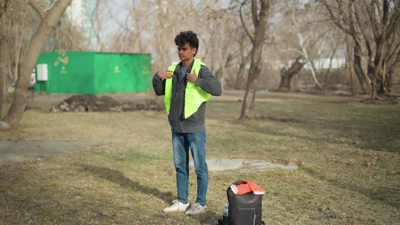 Man outdoors putting on bright neon green safety vest while standing in park with leafless trees, preparing for outdoor cleanup or volunteer work, backpack with gloves and water bottle placed on grass