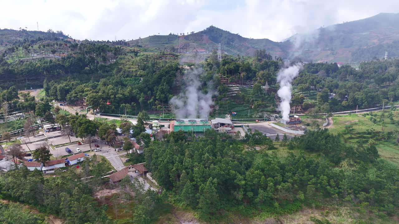 Aerial view of geothermal power plant operating amid lush forested hills, with visible steam vents releasing geothermal heat into the atmosphere. Dieng, Indonesia