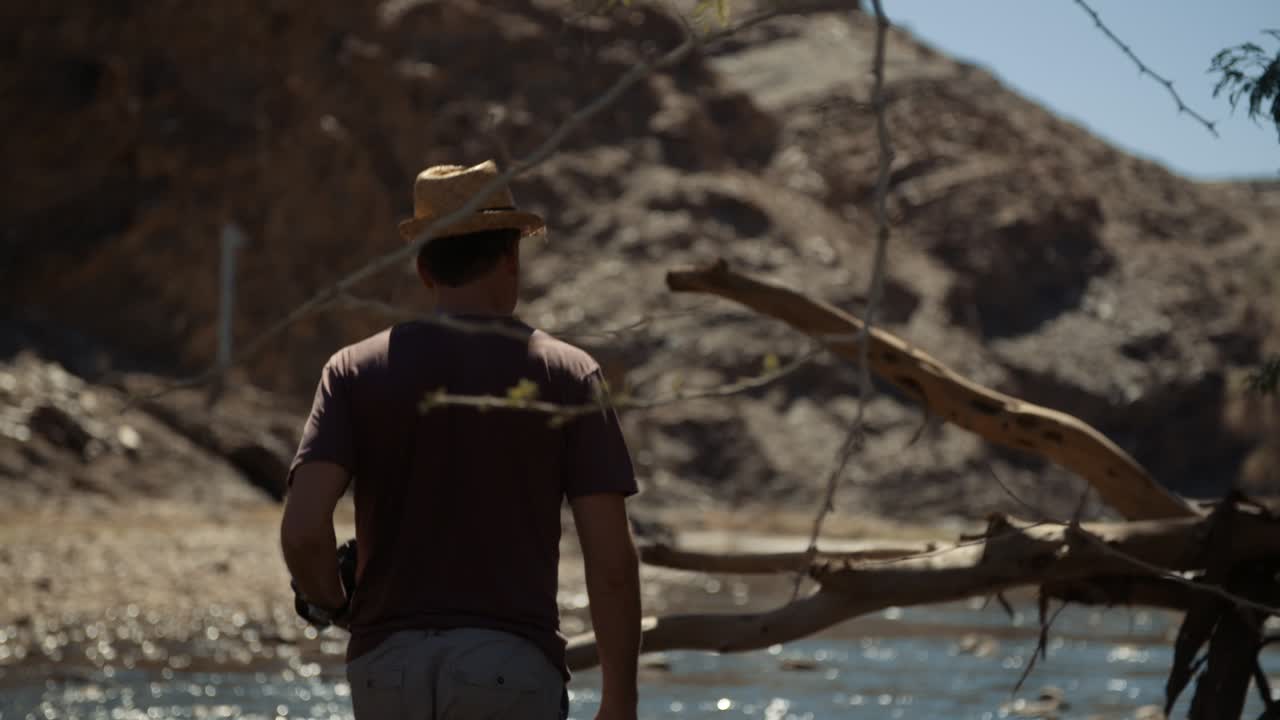 Handheld shot of a Caucasian photographer tourist with a stylish hat and camera walking through a river bed with a large tree stump in Africa and a mountain in the background.