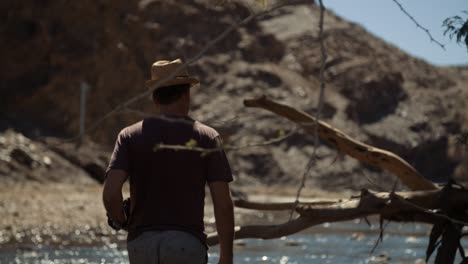 handheld shot of a caucasian photographer tourist with a stylish hat and camera walking through a river bed with a large tree stump in africa and a mountain in the background.