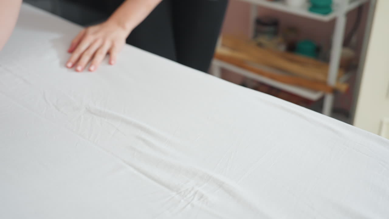 Light skin woman gently caressing white sheet on massage bed with both hands to remove wrinkles and ensure smoothness, capturing calm motion and soft detail in preparation for therapeutic use
