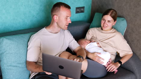 contemporary parenthood combining work. man shows something on laptop to his wife breastfeeding a baby.