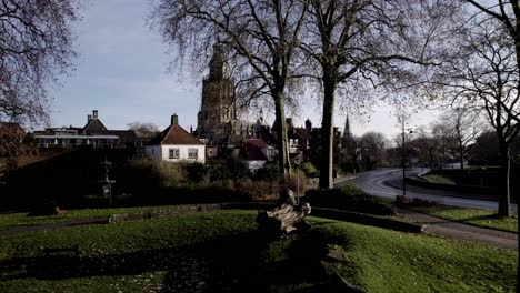 Descending-aerial-reveal-of-small-green-park-along-town-road-with-towerering-church-in-the-background-of-picturesque-city-seen-through-winter-barren-branches-of-a-tree