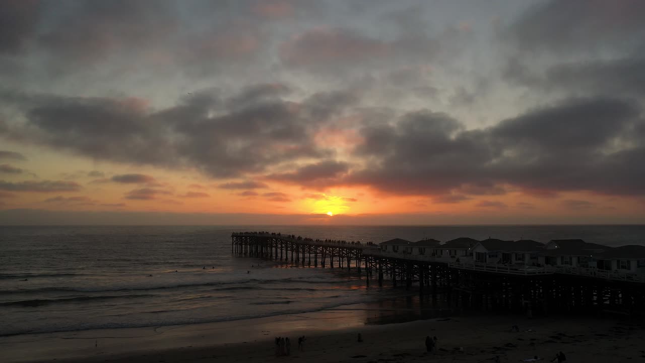 Drohne fliegt bei Sonnenuntergang nach links entlang des Crystal Pier in Pacific Beach, San Diego, und fängt Meereswellen und einen leuchtend orangefarbenen Himmel ein.