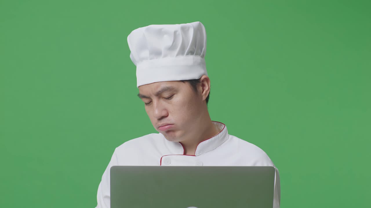 Close Up Of Asian Man Chef Using A Laptop And Having A Headache While Standing In The Green Screen Background Studio