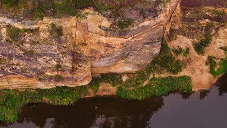 a detailed aerial view of erglu ergelu klintis, latvia, showcasing rugged sandstone cliffs, a calm river, and lush vegetation.