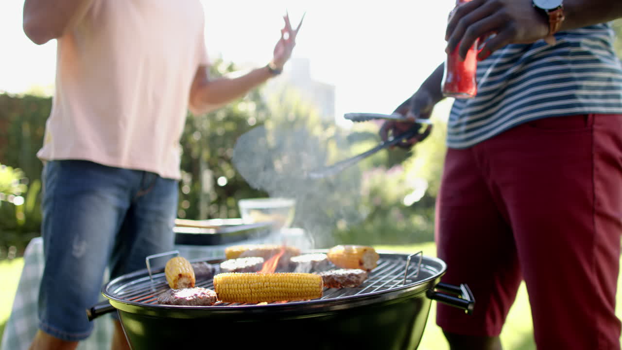 Grilling corn and burgers on barbecue grill, two multiracial male friends enjoying outdoor hangout