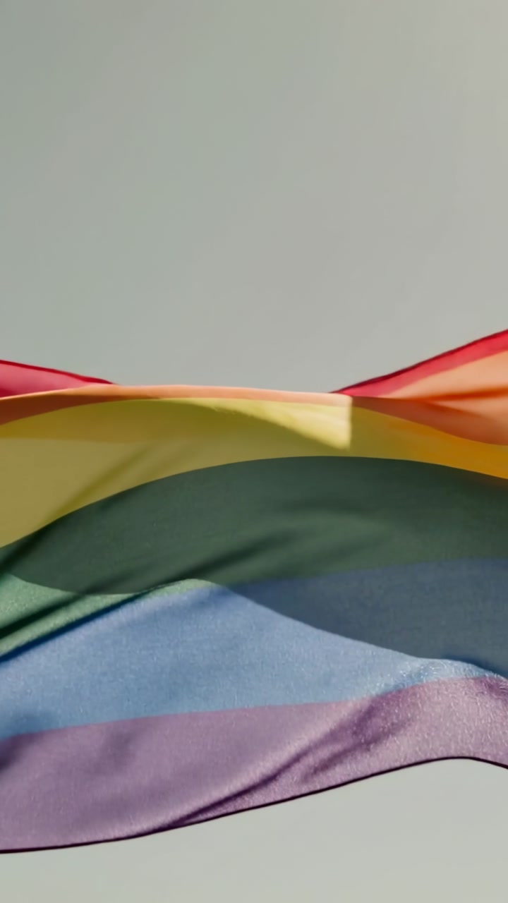 Vertical video: Waving rainbow flag rippling across pale sky, light breeze moving colored folds
