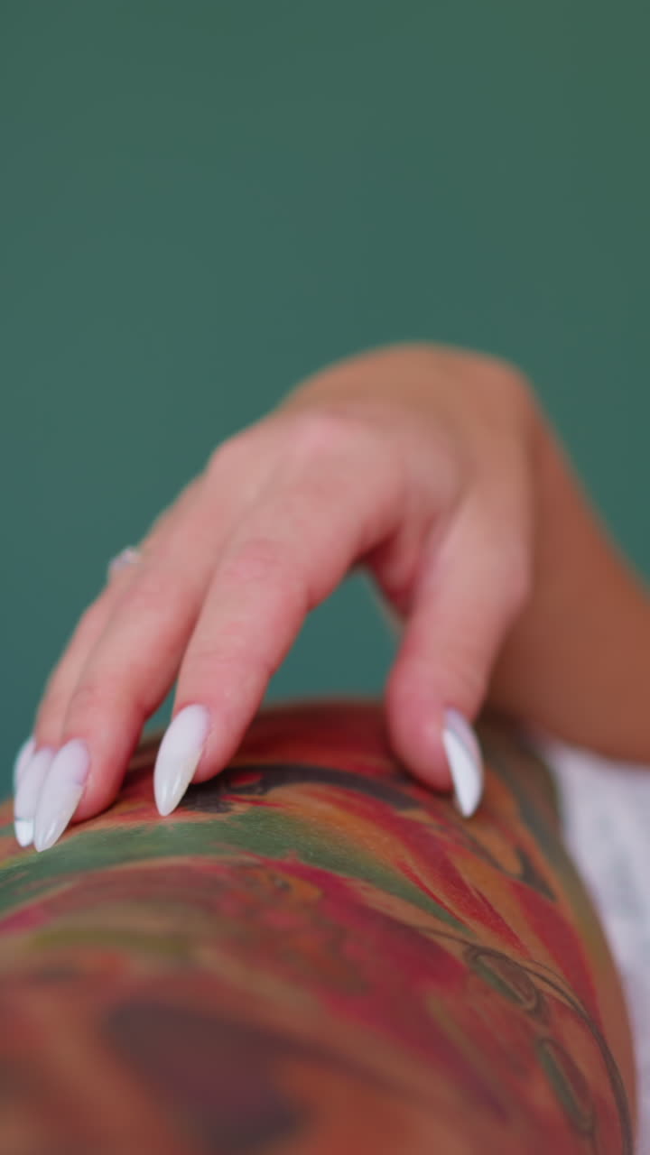 Seductive slim woman wearing white lacy lingerie holds hand on thigh covered with colorful tattoos pattern on green background closeup slow motion