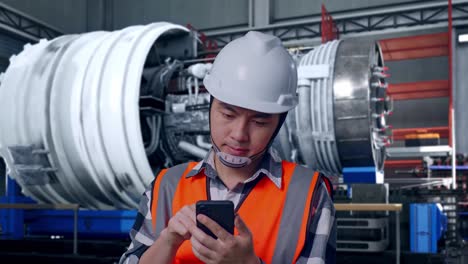 close up of asian male engineer with safety helmet using smartphone while standing with airplane engine maintenance conducted, analytics checking the turbine