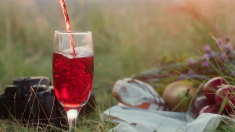 Close up of wine being poured into glass standing on grassy field beside camera, apples, pear, flowers, and cloth bag during outdoor picnic in warm natural light