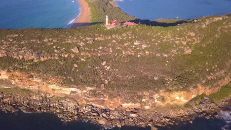 Toma-Aérea-Reveladora-Del-Faro-Principal-De-Barrenjoey-En-La-Parte-Superior-De-La-Roca-De-Arenisca-Del-Promontorio-Al-Atardecer-Del-Amanecer,-Volando-Sobre-La-Playa-De-Palmeras-Y-La-Bahía-Rota-En-La-Costa-De-Sydney-Al-Fondo