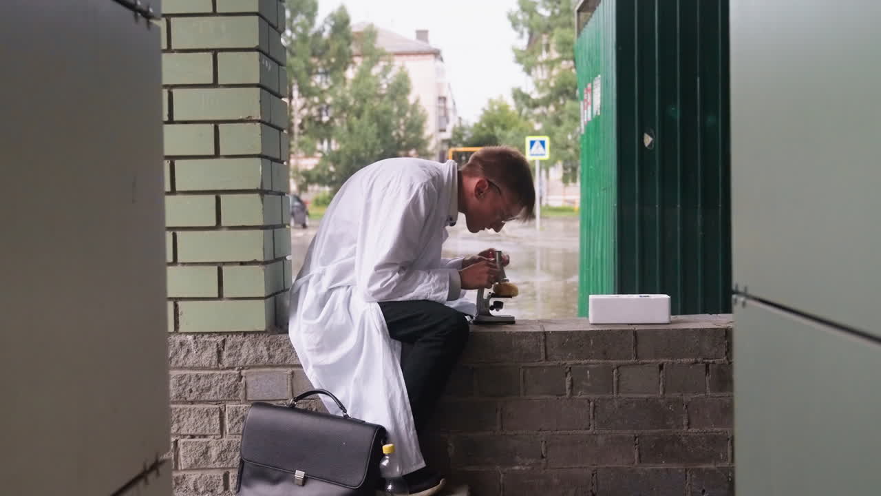 Young man in white coat sits outdoors carefully examining pastry with magnifying glass before observing through microscope, reflecting curiosity, scientific observation, and dedication