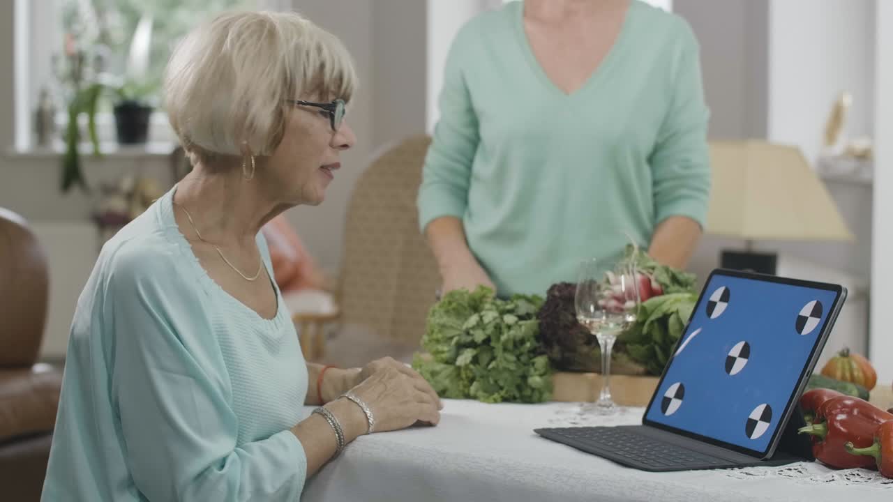 Side view of slim senior woman reading recipe from web-page on blue screen laptop as unrecognizable retiree cooking healthful food at background. Positive Caucasian friends indoors. Cinema 4k ProResHQ.