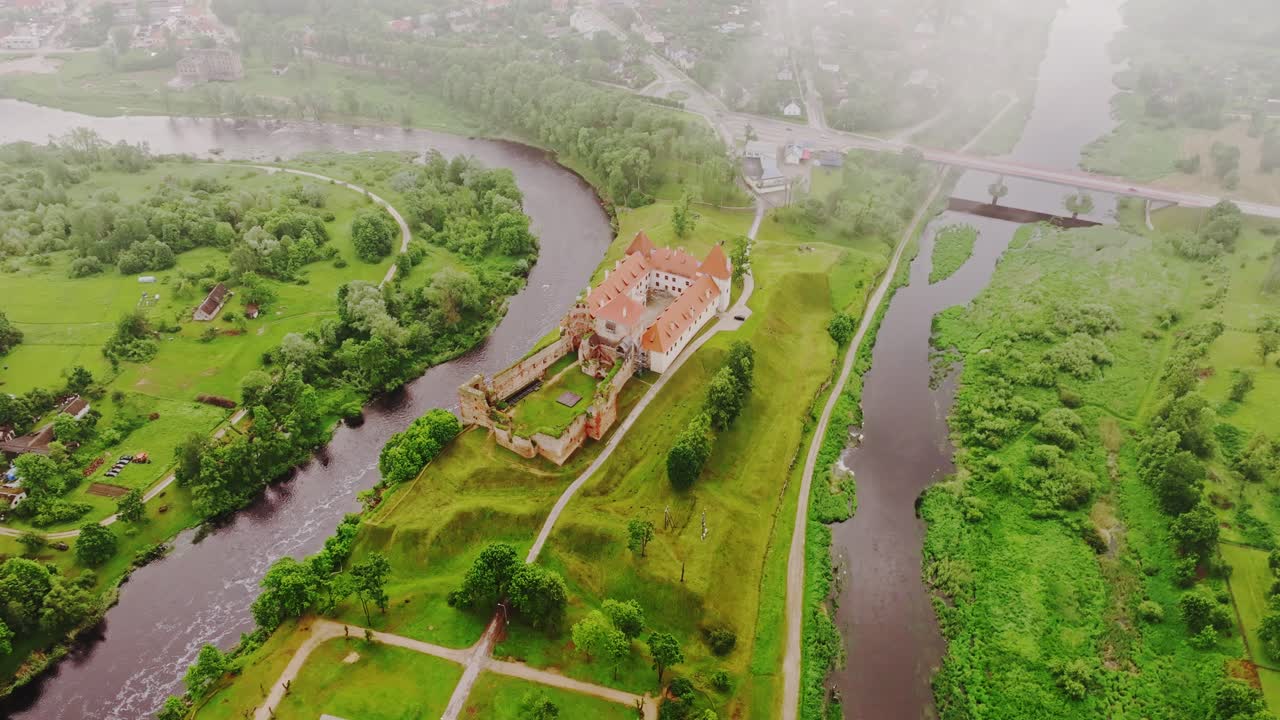 Drone slowly pulls away showing Bauska Castle between two rivers under clouds