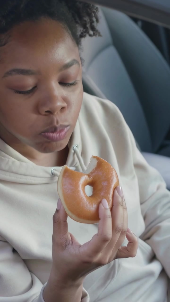 Vídeo vertical: Mulher de moletom com capuz pegando um donut, lanchando em um carro estacionado e verificando a hora no smartwatch.